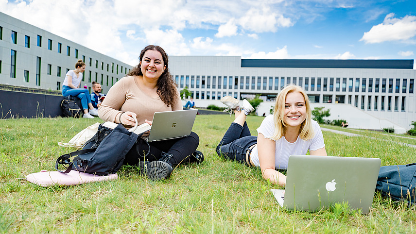 Two female students on a lawn in front of the library. One of them is sitting, the other is lying on the grass, both have laptops in front of them. Other students can be seen in the background.