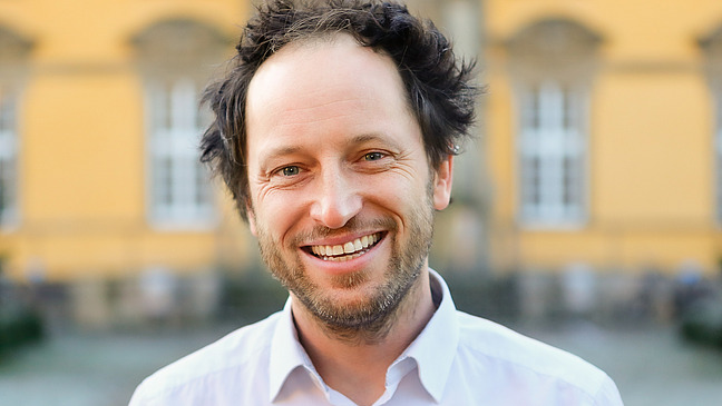 A smiling man with curly brown hair stands in front of a bright yellow façade. He is wearing a white shirt and has a friendly appearance.