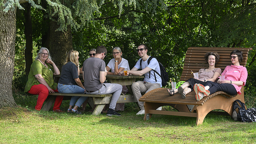 Mehrere fröhliche Personen mit Getränken bei einer Gartenparty, sechs sitzen an einem Tisch unter einem Baum, zwei liegen auf einer Liegebank.