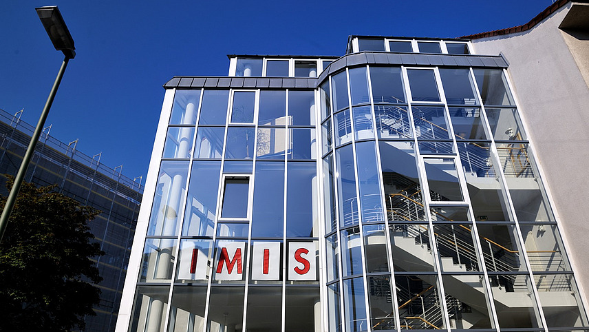 Glasfront eines Gebäudes an einem sonnigen Tag. Im zweiten Stockwerk hängen Plakate an der Glasfront, sodass von Außen "IMIS" zu lesen ist, was für das Institut für Migrationsforschung und Interkulturelle Studien steht. 