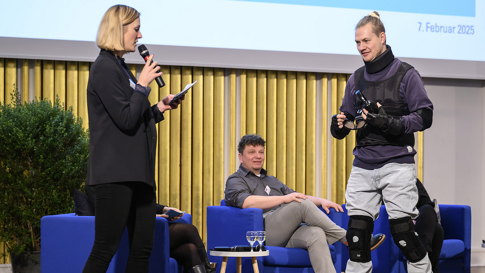 On stage, a woman interviews a man wearing an age simulation suit.