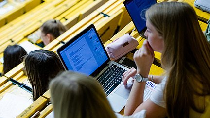 Zwei Studentinnen in einem Hörsaal. Ein von ihnen hat einen Laptop.