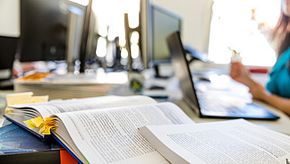 A table with several computer screens in the background. In the foreground is an open book with a bookmark and a colorful cover. Next to it are stacks of documents and a laptop. A person is partially visible while holding something in their hand.