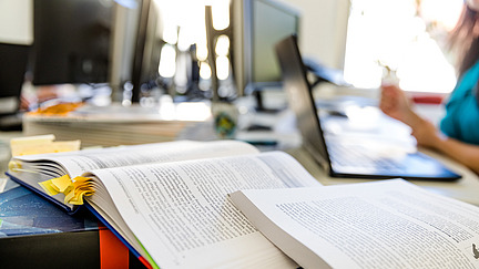 A table with several computer screens in the background. In the foreground is an open book with a bookmark and a colorful cover. Next to it are stacks of documents and a laptop. A person is partially visible while holding something in their hand.