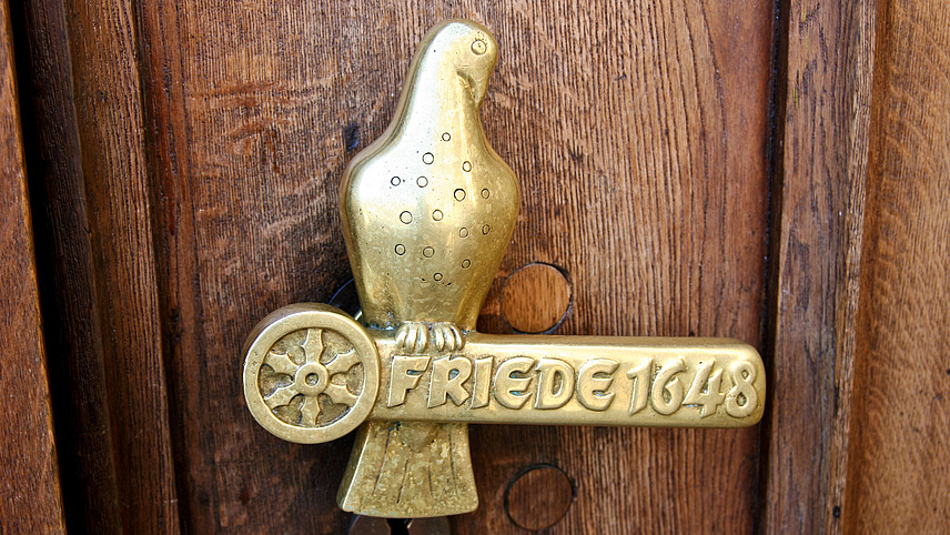 The picture shows an antique brass door fitting in the shape of a dove sitting on a text field. The text field bears the inscription “FRIEDE 1648”.