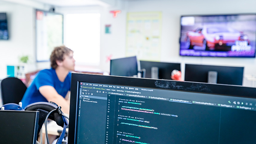 In front is a screen with a program code, while in the background a screen shows a car and a man looks focused at the screen in front of him.