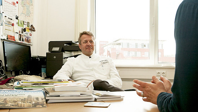 The picture shows a doctor in an office, sitting relaxed at a table and talking to another person whose hands are visibly gesticulating. The room is equipped with a computer, a printer and several stacks of documents and magazines