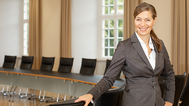 A woman in business attire stands in front of a large conference table and smiles at the camera