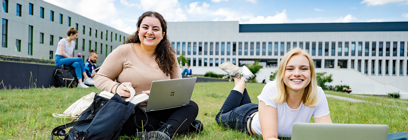 Two students on the grassy square next to the library. One of them is sitting, the other is lying on the grass, both have laptops in front of them.