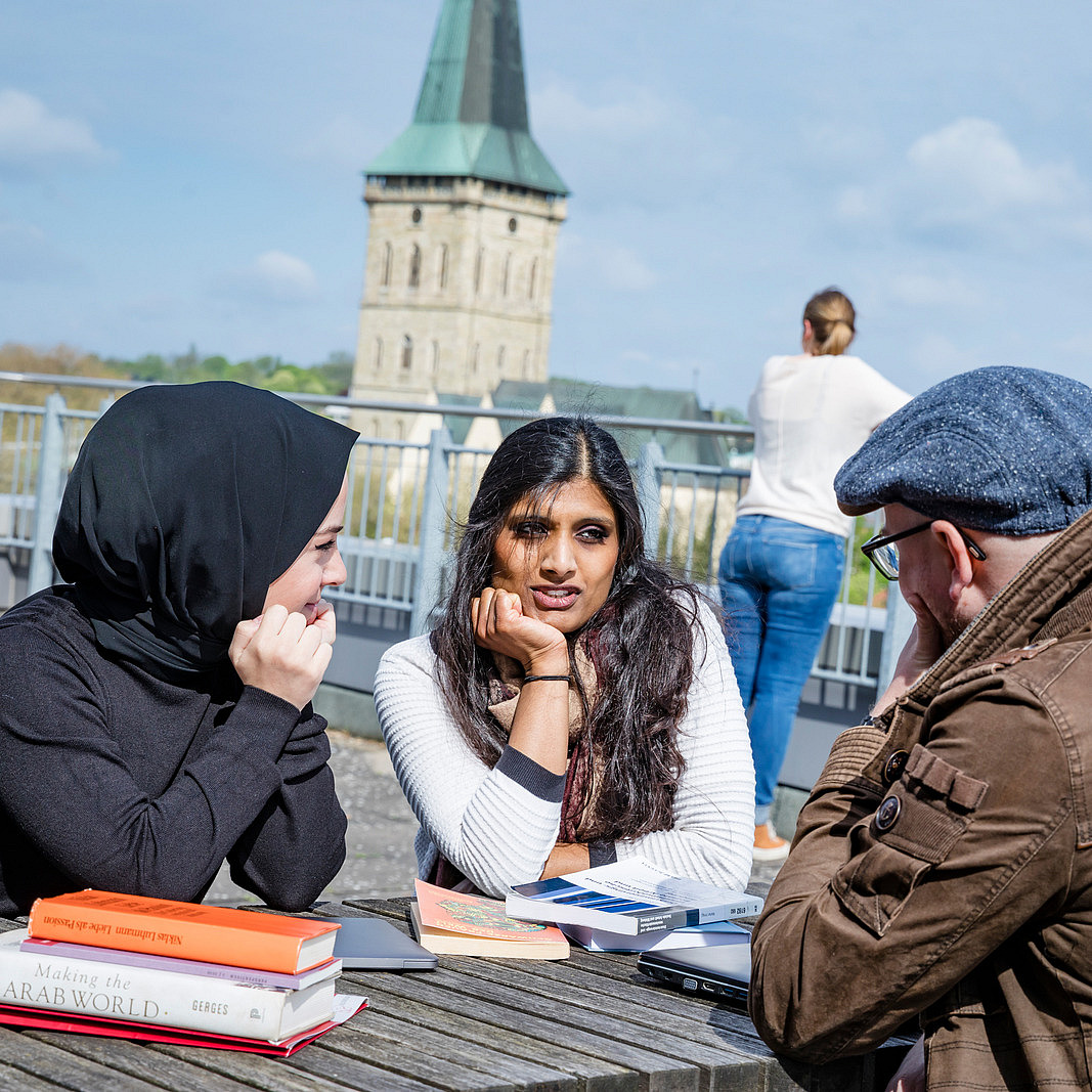 Drei Personen sitzen an einem Tisch im Freien. Eine von ihnen trägt ein Kopftuch. Vor ihnen liegen mehrere Bücher, eins hat den Titel "Making the Arab World". Im Hintergrund ist die Sankt Katharinen Kirche zu sehen.