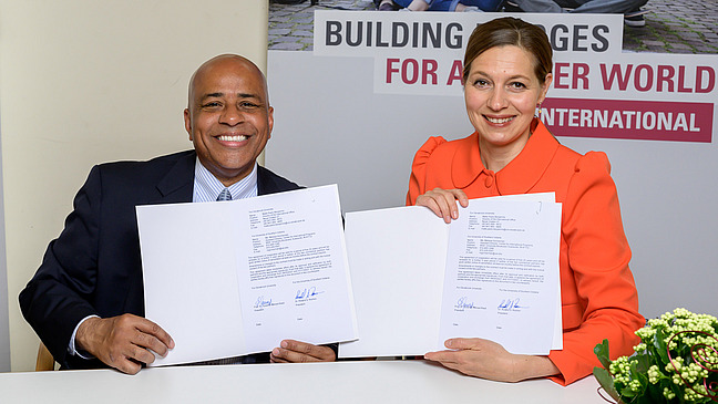 Prof. Dr. Susanne Menzel-Riedl and Dr. Roland S. Rochon pose for a photo in front of a stand with the title “Building Bridges for a better world. UOS international” while sitting for a photo. They are both holding a signed document in their hands and smiling at the camera.