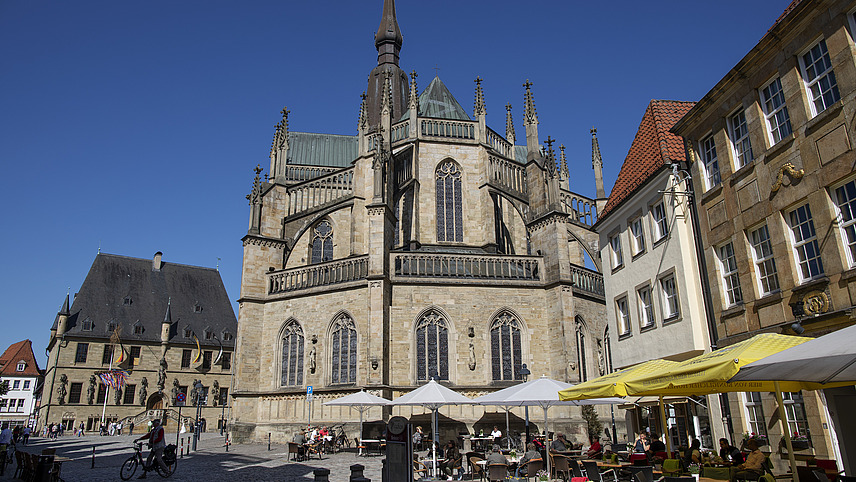 Der Osnabrücker Marktplatz mit Cafés im Vordergrund und mit dem historischen Rathaus und der evangelischen St. Marien Kirche im Hintergrund bei sonnigem Wetter.