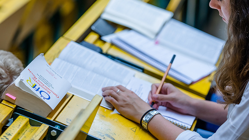 Eine Studentin macht sich Notizen und liest in einem Buch während einer Vorlesung. An ihrem Platz liegt ein weiteres Buch mit dem Titel: "Basistexte Öffentliches Recht".