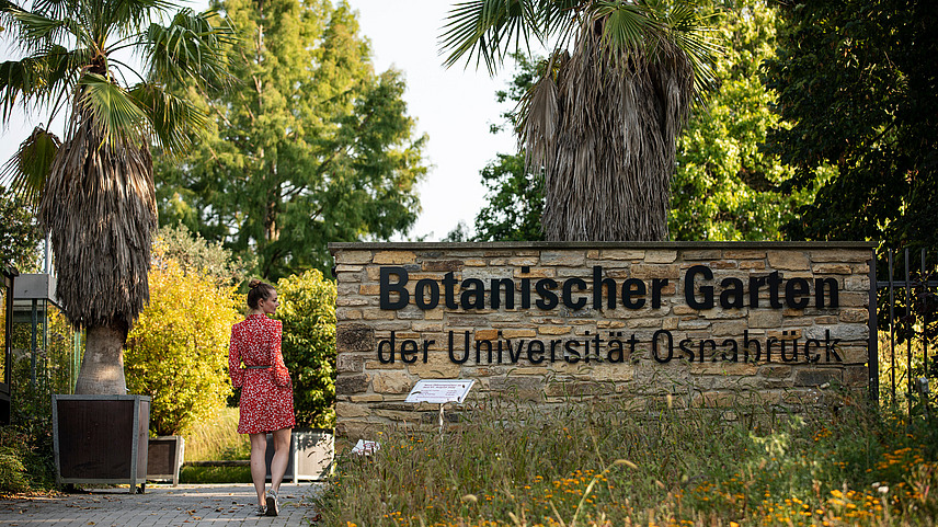 Eine Frau schreitet durch den neuen Eingang des botanischen Gartens. Als großer Schriftzug auf einer Mauer steht "Botanischer Garten der Universität Osnabrück".