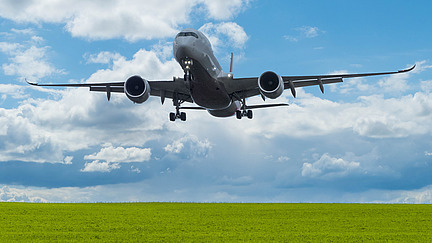 An airplane flies over a green meadow under a blue sky with a few clouds.