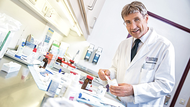 A man in a white lab coat stands at a table covered with various medicines and packaging. He smiles in a friendly manner and points to a product while conveying a professional atmosphere.