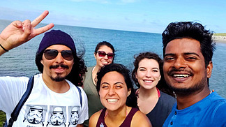 Five international students stand smiling in front of the sea. It is sunny and they are all wearing light clothing.