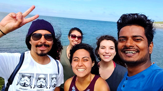 Five international students stand smiling in front of the sea. It is sunny and they are all wearing light clothing.