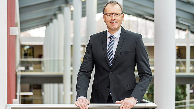 The image shows a man in a black suit and a striped tie, standing and smiling while leaning on a railing in a modern architectural setting with large columns and a bright interior. 