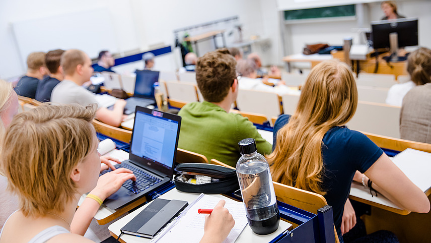 Studierende sitzen im Hörsaal während einer Vorlesung und machen sich Notizen oder tippen am Laptop.