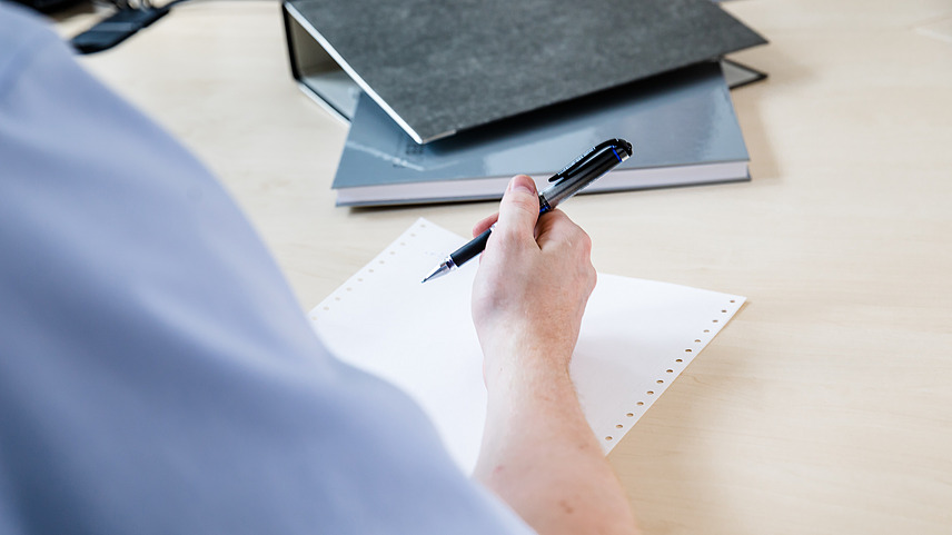 View over the shoulder of a person dressed in a shirt, writing on a sheet of paper with a ballpoint pen.