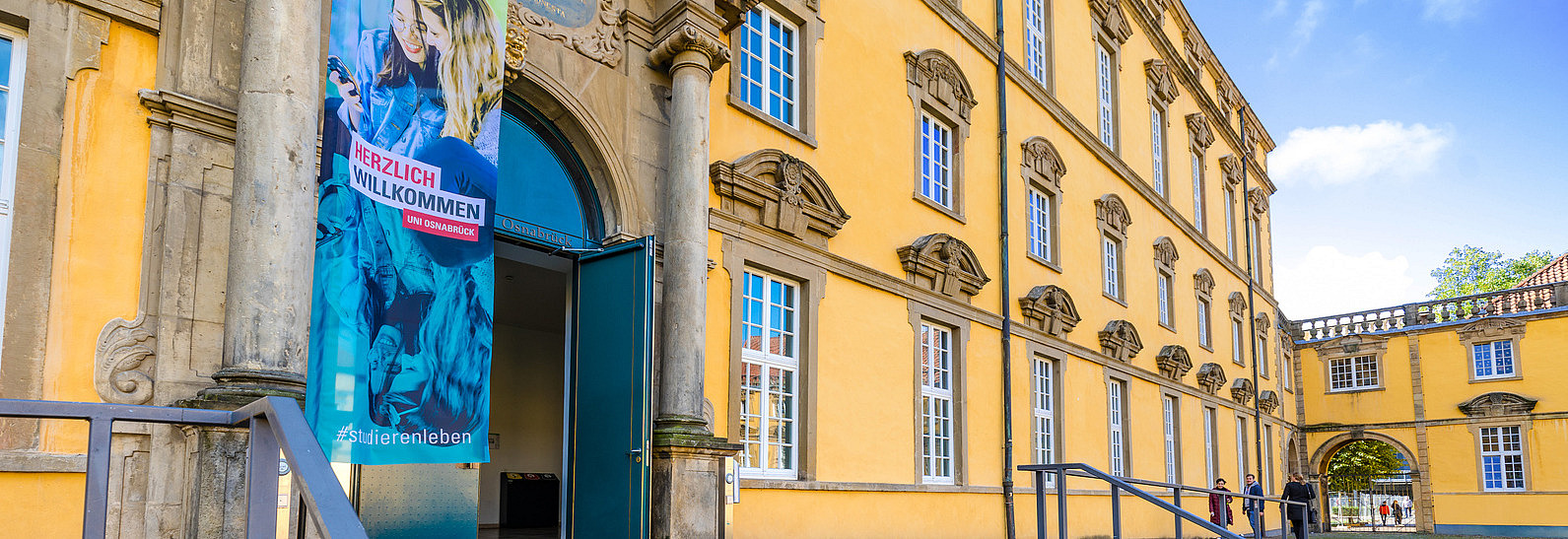 Der Eingang zu einem historischen Gebäude ist im Bild sichtbar, mit einer einladenden Tür und einem großen Banner, das "Herzlich Willkommen" zeigt. Vor den Stufen liegt ein weiterer Willkommensplakat mit der Aufschrift "SCHÖN, DASS DU DA BIST!". Die Fassade des Gebäudes ist in warmen Gelbtönen gehalten.