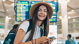 A young woman stands smiling in summer clothes with a hat on her head, backpack on her shoulders and cell phone in her hand in an airport hall. In the background are digital boards with exhaust air information.