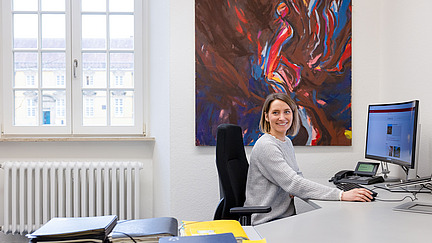 A woman sits at her desk in an office and looks into the camera with a smile. Folders and notepads in bright colors lie on the table. A large work of art can be seen on the wall in the background. The room looks bright and friendly.