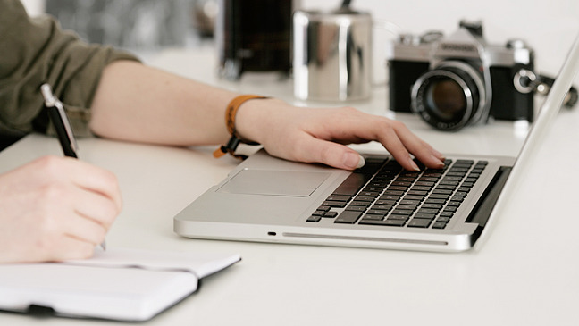 Woman's hand on laptop keyboard, the other hand writing in a notebook with a ballpoint pen, camera in the background