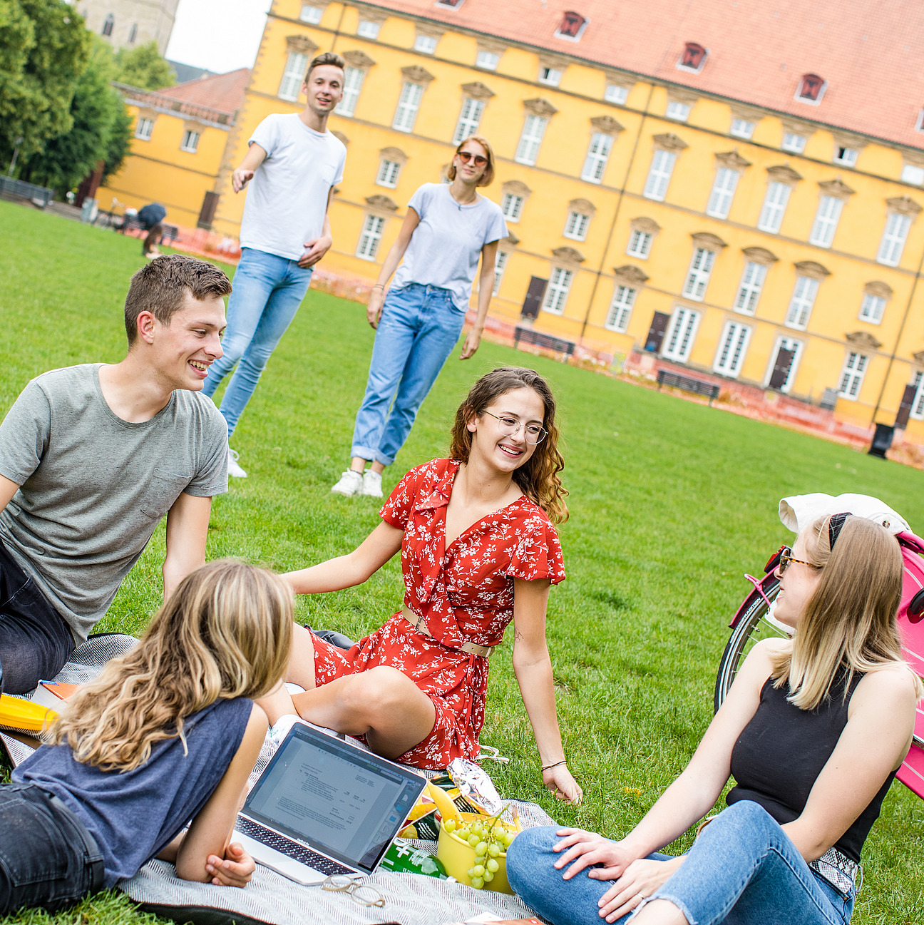 Vier Studierende sitzen auf einer Decke im Schlossgarten. Zwei weitere gehen auf sie zu. Neben ihnen steht ein pinkes Fahrrad. Im Hintergrund ist das Schloss zu sehen.