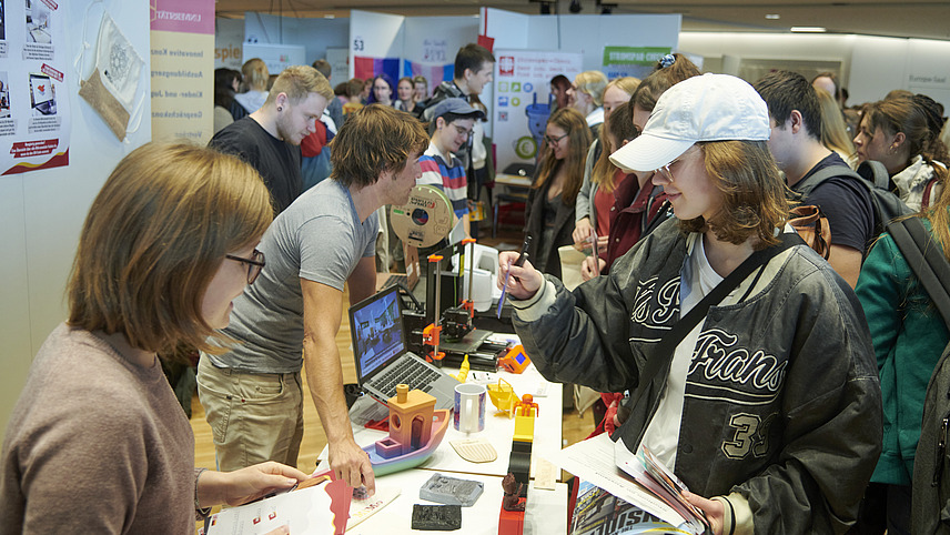 There are many different information stands in a hall, which are well attended by students. On the table of the information stand in the foreground you can see various utensils such as a model of a ship and small figures printed with a 3D printer.