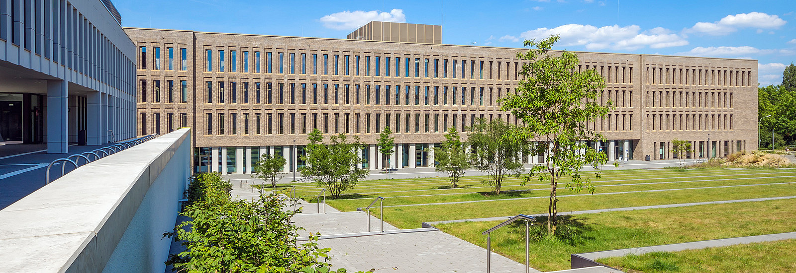 A modern building front can be seen in the picture, which is made of dark bricks and has large windows. In the foreground is a green area with lawns and small trees. Stone steps lead through the outdoor area, which is complemented by inviting architecture. 