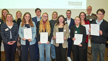 A group of several people stands smiling in front of a wall decorated with a sign reading "Deutschland Stipendium." Everyone is holding certificates in their hands. 