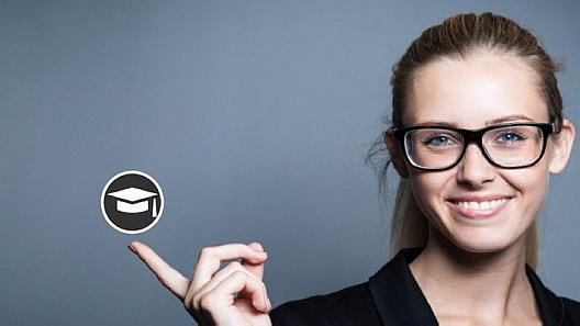 Smiling woman with glasses, dressed in a formal outfit. She holds out her index finger and points to a black symbol with a graduation hat, which is shown floating in the air.