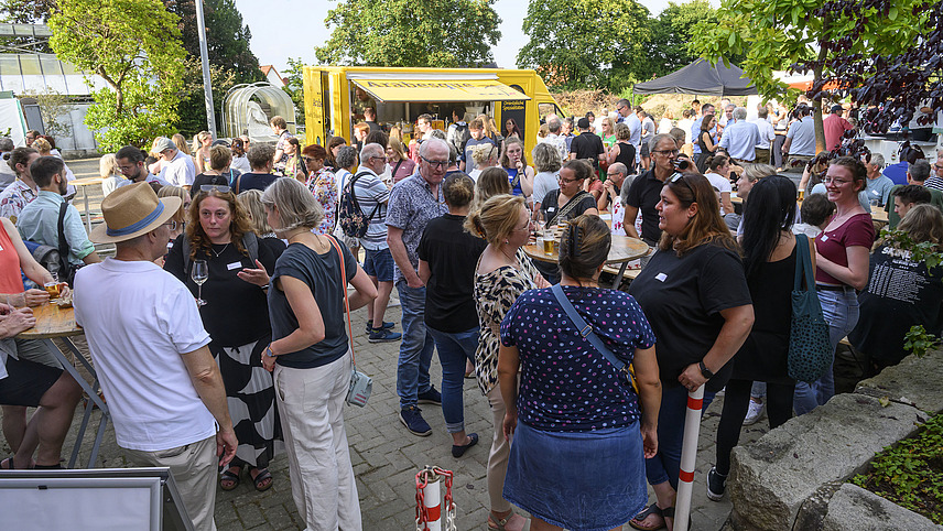 A crowd at a garden party is standing in front of food stands.