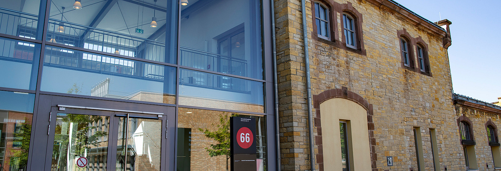 Eine Kombination aus modernem und historischem Design ist zu sehen. Im Vordergrund befindet sich ein Glasbereich mit doppelten Türen und einer großen Anzeige mit der Nummer 66. Neben dem modernen Eingang ist eine Fassade aus Sandstein sichtbar.