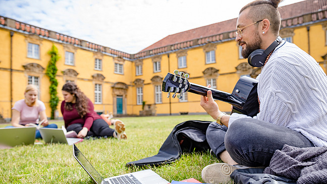 Three students are sitting together in the castle courtyard. They have laptops and books lying next to them. One of them is playing the guitar.