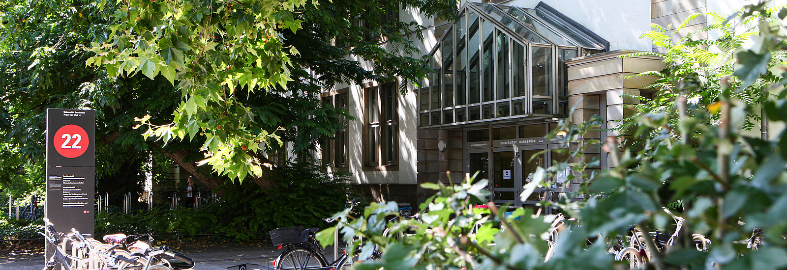 An entrance to a building is surrounded by trees and plants. Numerous bicycles are parked in the foreground, while a sign with the number 22 is visible. The sun's rays shine through the foliage in the A view of a historic building with a red pitched roof and large windows can be seen in the foreground. It is surrounded by trees.