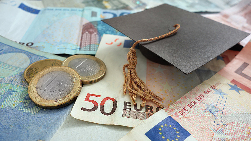 A small cardboard doctor's hat lies on 50 and 20 euro bills and next to it are a few individual euro coins.