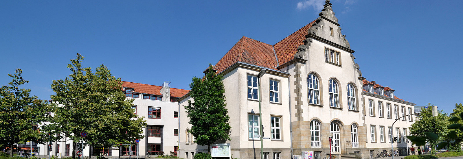 A view of a historic building with a red pitched roof and large windows can be seen in the foreground. It is surrounded by trees, while in the background there are modern buildings and a clear blue sky.