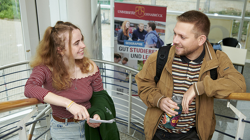 A woman and a man stand laughing in a stairwell. In the background is a banner with the word “Geography” printed on it.