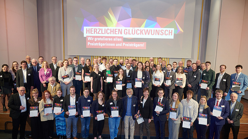 Eine Gruppe von Menschen steht auf einer Bühne und hält Urkunden in den Händen. Die Personen lächeln und sind in formeller Kleidung.