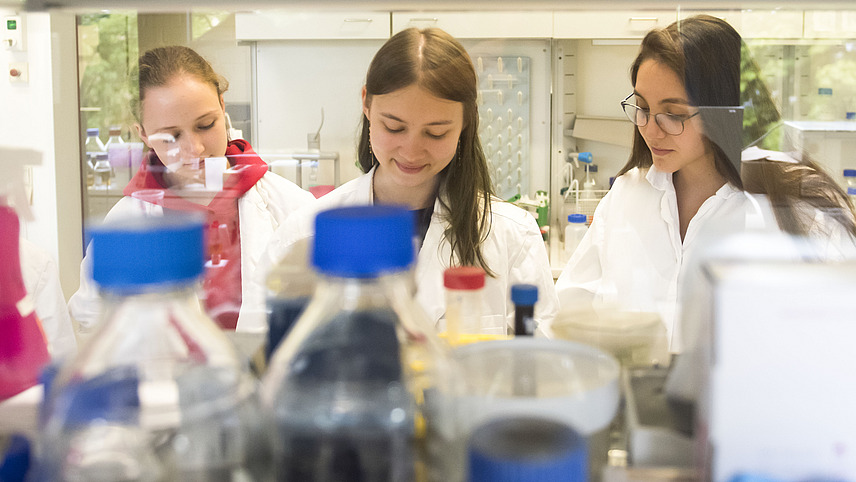 Three students in lab coats stand behind a shelf of chemicalsShelf 