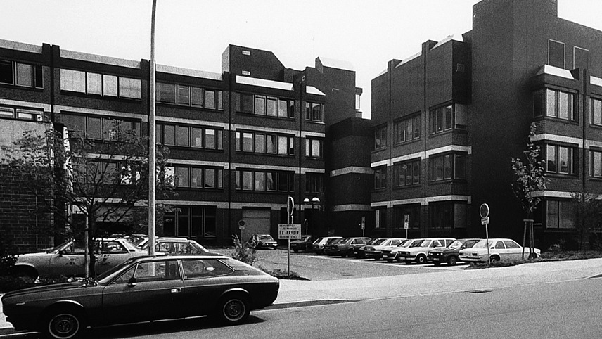 Schwarzweiß-Aufnahme des Gebäudes, davor ein Parkplatz mit Fahrzegen aus den 1970er und 1980er Jahren