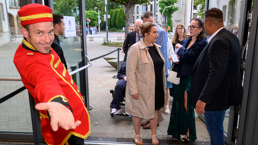 A person in a bellboy costume looks into the camera with a welcoming gesture