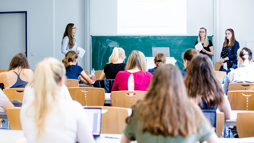 Blick von hinten über die Sitzreihen eines Seminarraums. Doktor Monika Voges leitet das Seminar und steht vorne an der Tafel neben zwei Studentinnen.