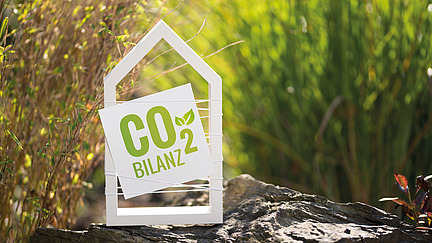 Ein Schild in Form eines Hauses zeigt die Aufschrift "CO₂-BILANZ" in grüner Schrift. Es steht auf einem Felsen, umgeben von grünen Pflanzen und Gras im Hintergrund. Der Fokus auf dem Schild betont das Thema Umweltbewusstsein und Nachhaltigkeit.