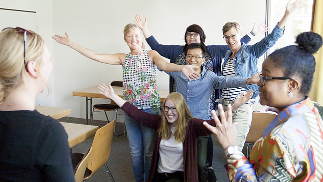 A group of seven people pose happily in a room. They stand in various poses and radiate joy as they raise their arms.