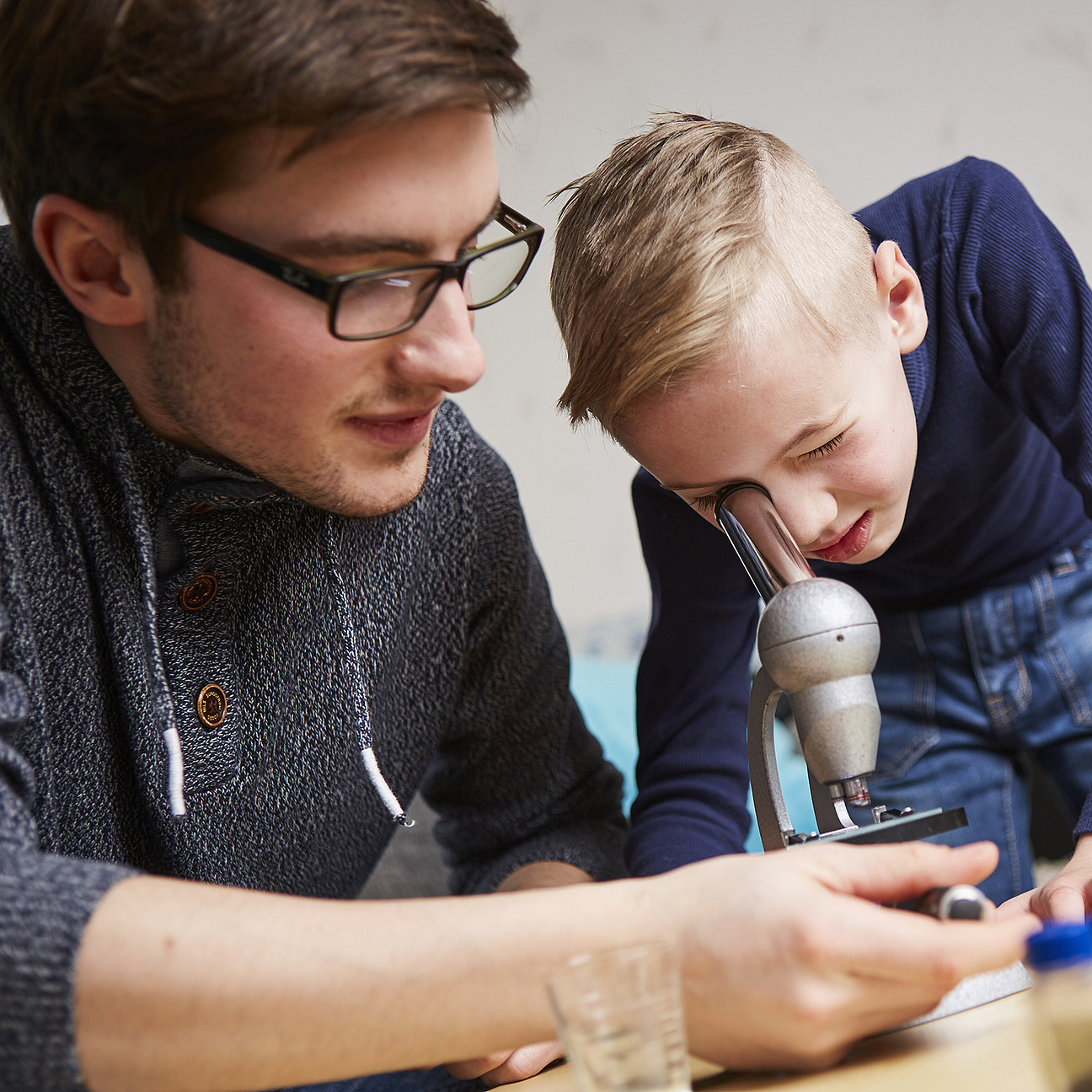 A young man wearing glasses and a gray sweater sits next to a small boy who is curiously looking into a microscope. The boy has short blonde hair and is focused. The man points at the microscope with his hand, likely explaining something.