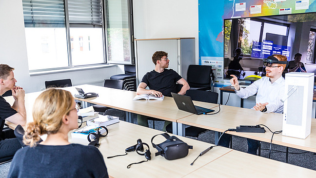 Jannis Vogel hat eine augmented reality Brille auf und greift mit seiner Hand nach etwas. Hinter ihm ist seine Sicht auf einen Bildschirm übertragen. Er navigiert gerade ein Menü.